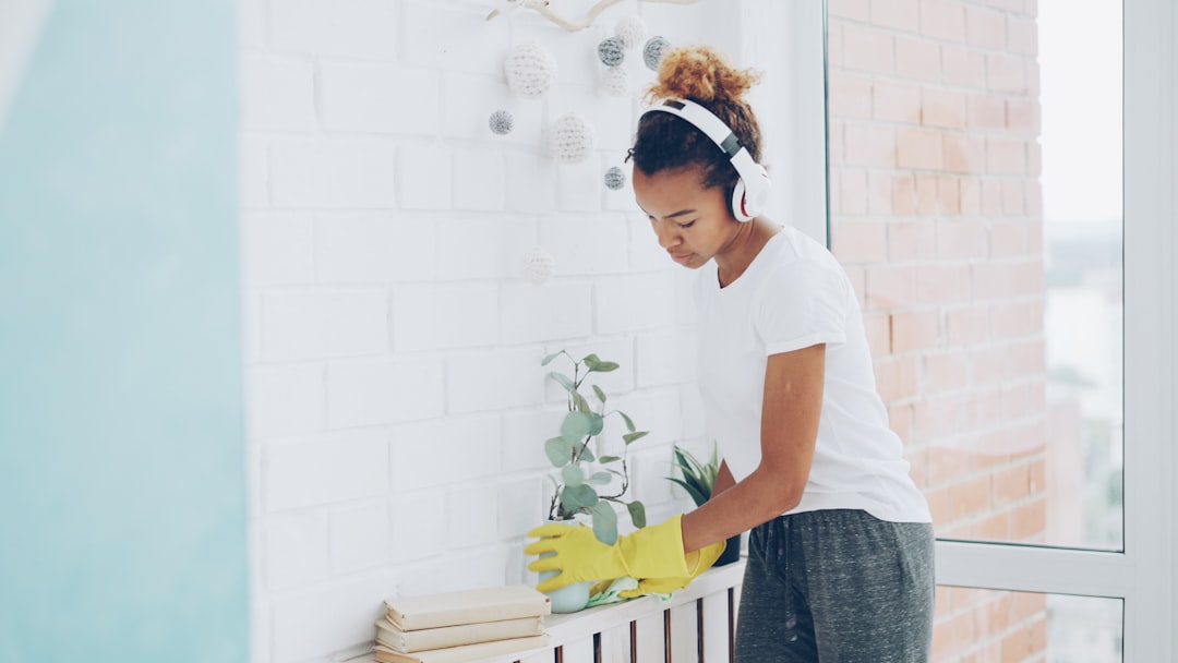 Cheerful maid African American girl is doing housework dusting using cloth and wearing protective gloves, young woman is listening to music in headphones