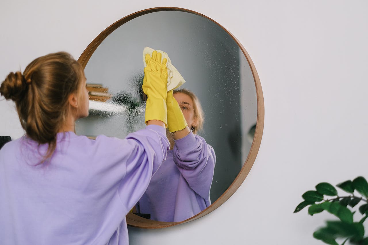 about-us A woman wearing yellow gloves cleans a round wall mirror indoors, reflecting her image.